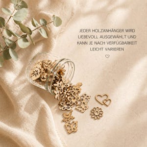 May include: A clear glass jar tipped on its side, spilling small, light-brown wooden cutouts in shapes of butterflies, flowers, birds, and hearts. The text JEDER HOLZANH&Auml;NGER WIRD LIEBEVOLL AUSGEW&Auml;HLT UND KANN JE NACH VERF&Uuml;GBARKEIT LEICHT VARIIEREN is visible.