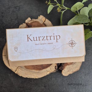 Könnte beinhalten: Ein rechteckiger beigefarbener Papierumschlag mit dem braunen Schriftzug "Kurztrip" und "ZEIT STATT ZEUG", einer gepunkteten Reiseroutenlinie, einem Standort-Pin-Symbol und einer Kompassrosengrafik auf der Vorderseite des Briefumschlags.