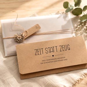 Pu&ograve; includere: Una busta rettangolare in carta marrone con il testo stampato in nero ZEIT STATT ZEUG, un piccolo cuore e la frase WEIL GEMEINSAME MOMENTE DIE SCH&Ouml;NSTEN GESCHENKE SIND. &Egrave; legata con uno spago sottile e una decorazione a forma di fiore in legno.