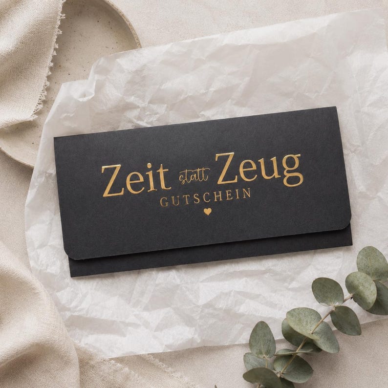 May include: A rectangular black envelope featuring gold foil lettering that reads Zeit statt Zeug GUTSCHEIN with a small gold heart icon centered below the text. The envelope is placed on a piece of white crinkled tissue paper.