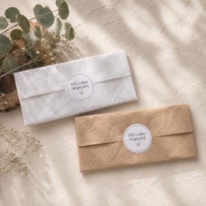 May include: Two rectangular packages wrapped in paper, one white and one brown, each sealed with a circular sticker featuring the text Mit liebe verpackt and a small heart icon. The packages are arranged side by side on a textured surface.