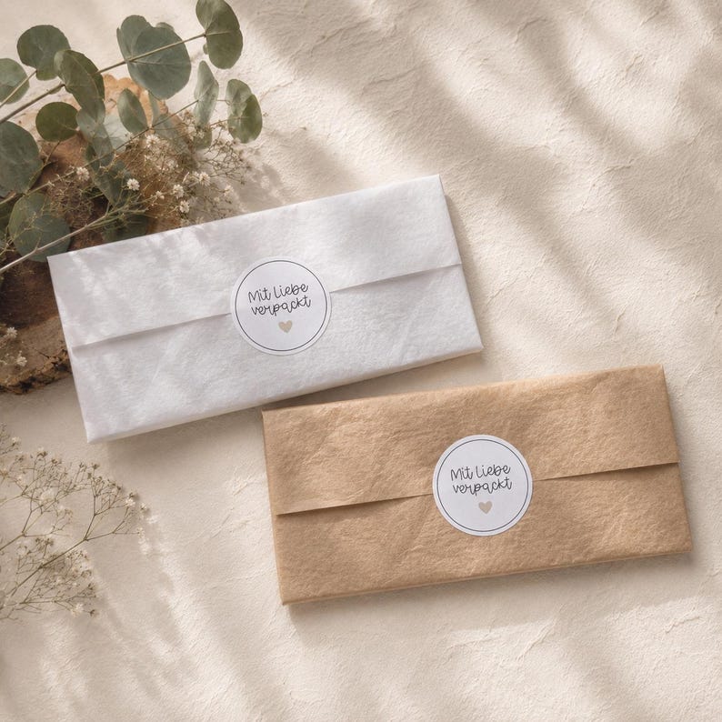May include: Two rectangular packages wrapped in paper, one white and one brown, each sealed with a circular sticker featuring the text Mit liebe verpackt and a small heart icon. The packages are arranged side by side on a textured surface.