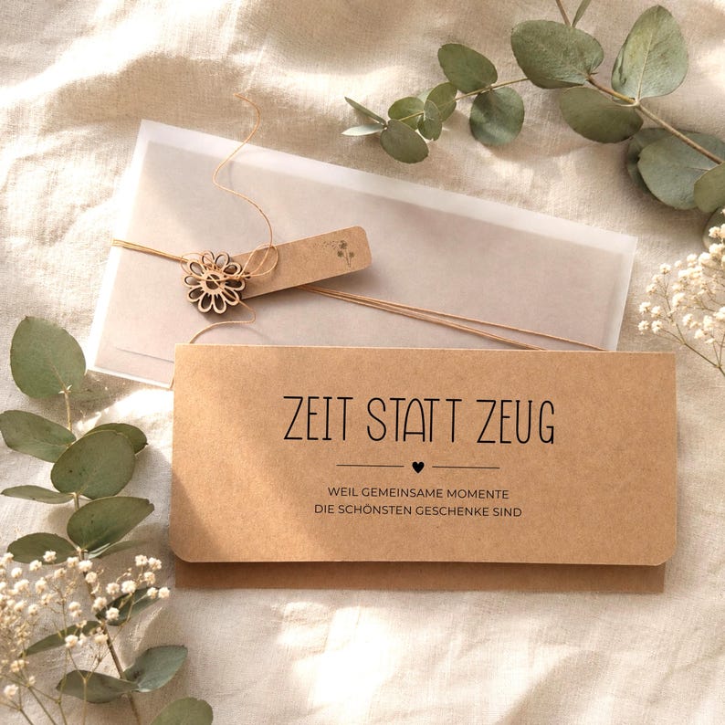 Pu&ograve; includere: Una busta rettangolare in carta kraft con il testo tedesco ZEIT STATT ZEUG e un sottotitolo, decorata con un ciondolo in legno a forma di fiore legato con uno spago sottile, appoggiata su una busta traslucida e circondata da foglie di eucalipto.