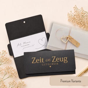 May include: A black envelope containing a white card with the text "f&uuml;r Mama" and a heart illustration, alongside a second black envelope with gold text "Zeit statt Zeug GUTSCHEIN" and a heart icon, plus a translucent envelope with a heart charm.