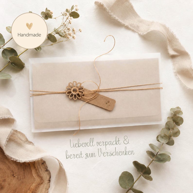 May include: A rectangular envelope in a translucent white sleeve, tied with thin tan twine and a small wooden flower cutout. A rectangular kraft paper tag is attached to the twine. The text "Liebevoll verpackt & bereit zum Verschenken" is printed below.