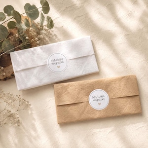 Pu&ograve; includere: Due pacchetti rettangolari avvolti nella carta, uno bianco e uno in carta kraft marrone. Ogni pacchetto &egrave; sigillato con un adesivo circolare bianco che riporta il testo Mit liebe verpackt e una piccola icona a forma di cuore al centro.