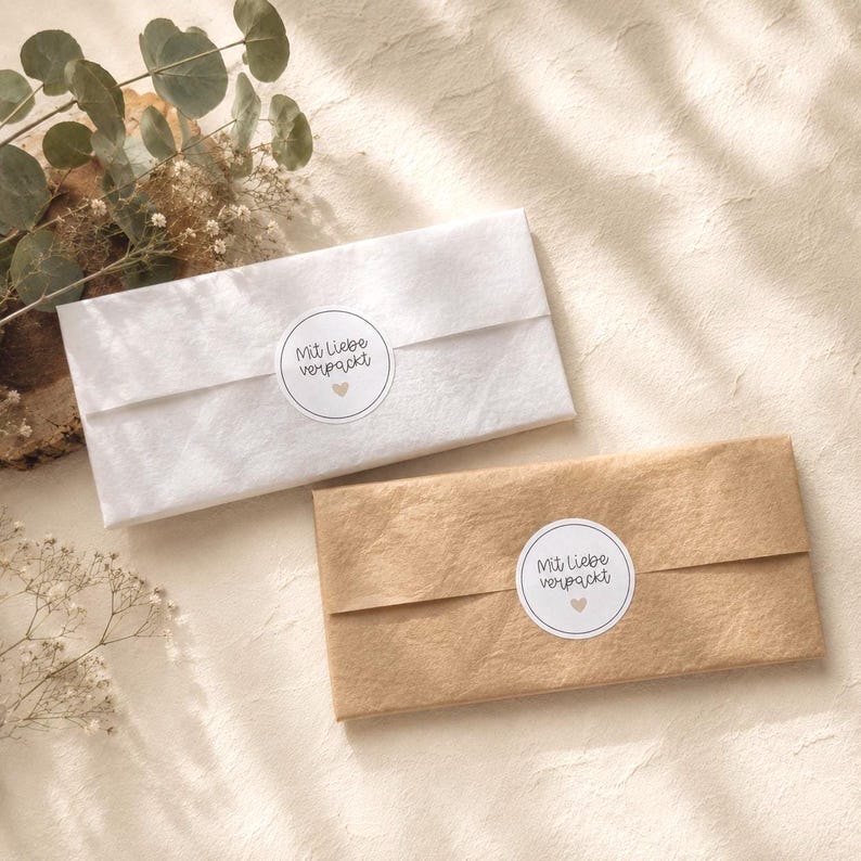 Pu&ograve; includere: Due pacchetti rettangolari avvolti nella carta, uno bianco e uno in carta kraft marrone. Ogni pacchetto &egrave; sigillato con un adesivo circolare bianco che riporta il testo Mit liebe verpackt e una piccola icona a forma di cuore al centro.