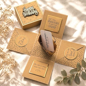 Könnte beinhalten: Eine braune, quadratische, handgefertigte Geburtstagsgeschenkbox mit einem Deckel mit der Aufschrift "Happy Birthday". Die Box öffnet sich und zeigt mehrere Tafeln mit Punkt- und Linienmustern. Jede Tafel hat eine Nachricht auf Deutsch.