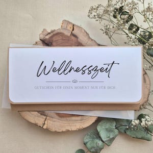 Könnte beinhalten: Ein rechteckiger Gutschein mit weißer Vorderseite und brauner Kraftpapier-Rückseite. Der Text lautet Wellnesszeit und GUTSCHEIN FÜR EINEN MOMENT NUR FÜR DICH, mit einem kleinen Lotusblüten-Symbol mittig zwischen den beiden Textzeilen.
