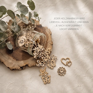 May include: A clear glass jar spilling out small, laser-cut wooden shapes including butterflies, flowers, birds, and hearts. The text JEDER HOLZANH&Auml;NGER WIRD LIEBEVOLL AUSGEW&Auml;HLT UND KANN - JE NACH VERF&Uuml;GBARKEIT - LEICHT VARIIEREN is printed on the right side.