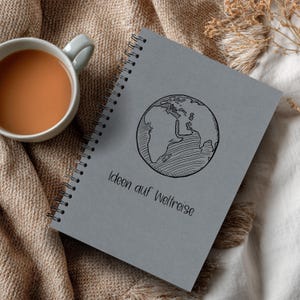 Könnte beinhalten: Ein graues Spiralnotizbuch mit einer schwarz-weißen Weltkugel-Illustration und dem Text "Ideen auf Weltreise". Links neben dem Notizbuch steht eine Tasse Tee auf einem beigen Stoff.