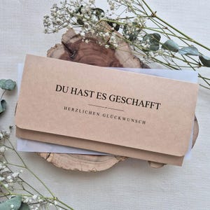 Könnte beinhalten: Ein rechteckiger hellbrauner Papierumschlag mit abgerundeten Ecken, der den schwarzen Aufdruck DU HAST ES GESCHAFFT und HERZLICHEN GLÜCKWUNSCH sowie eine horizontale Trennlinie zwischen den beiden Textzeilen aufweist.