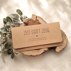 Pu&ograve; includere: Una busta rettangolare in carta kraft con il testo stampato in nero ZEIT STATT ZEUG, una piccola icona a forma di cuore e la frase tedesca WEIL GEMEINSAME MOMENTE DIE SCH&Ouml;NSTEN GESCHENKE SIND, appoggiata su una fetta di legno con eucalipto.