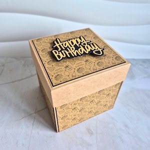 Könnte beinhalten: Eine quadratische, zweifarbige Geschenkbox mit Deckel mit den Worten "Happy Birthday" in Schwarz und Gold. Die Box ist beige mit einem sich wiederholenden Muster von Geburtstags-Symbolen. Der Deckel ist beige mit schwarzem Rand.