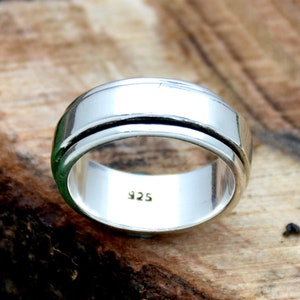 May include: A silver spinning ring with a 925 stamp on the inside. The ring is on a wooden surface.