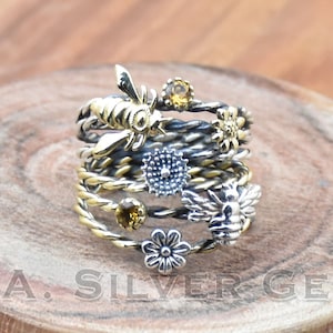 May include: A silver and gold ring with a bee, flower, and gemstone design. The ring is made of twisted wire and has a rustic look.