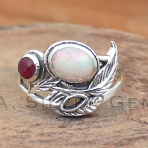 May include: A silver ring featuring an oval opal and a small, round ruby. The ring has a leaf design on the band and around the stones. The ring is set on a wooden surface.