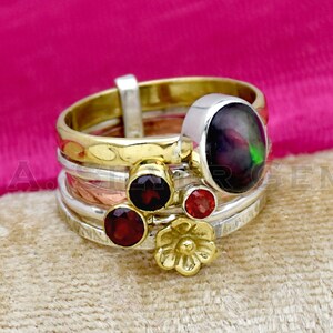 May include: A multi-layered ring with a black opal gemstone and red garnet accents. The ring is made of silver, gold, and copper.