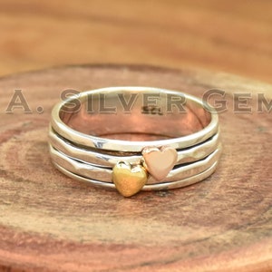Puede incluir: Un anillo de plata con tres bandas y dos adornos en forma de corazón, uno dorado y otro de oro rosa. El anillo está sobre una superficie de madera, y el texto "S. A. SILVER GEMS" es visible.