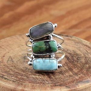 Pode incluir: Um anel de prata com três pedras retangulares empilhadas. A pedra superior é uma labradorita de cor púrpura escura, a pedra do meio é uma turquesa verde e a pedra inferior é uma turquesa azul céu.