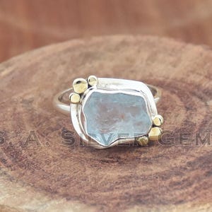 May include: A silver ring with a light blue, irregularly shaped gemstone. The ring has gold accents and is set on a wooden surface. The ring is a piece of jewelry.