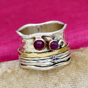 Peut inclure: Bague rotative argentée avec une bande texturée et deux petites pierres précieuses rondes rouge foncé. La bague a une bande dorée et une texture martelée sur la partie supérieure.