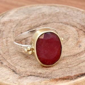 May include: A silver ring with a gold setting and a large, oval-shaped, red gemstone. The ring has a textured band and small gold accents.
