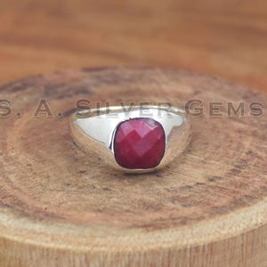 May include: A silver ring with a large, square-cut, deep red gemstone. The ring has a wide band and a polished finish. The text "S. A. SILVER GEMS" is visible on the wooden surface. This ring is a piece of jewelry.