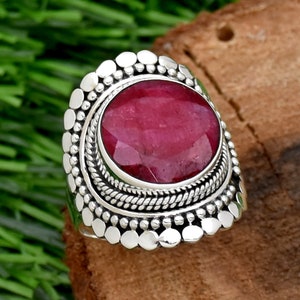 May include: A silver ring with a large, oval-shaped, deep red ruby gemstone set in a decorative, beaded bezel.