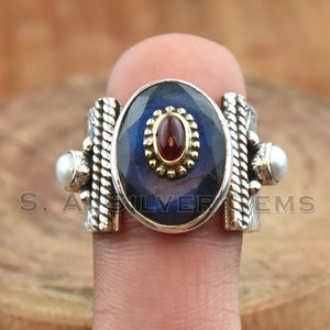 May include: A silver ring with a blue gemstone, a red gemstone, and two white pearls. The ring has a braided design on the sides.