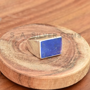Pode incluir: Um anel de prata com uma pedra retangular de lapis-lazúli azul. O anel está colocado sobre uma superfície de madeira.