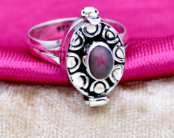Puede incluir: Un anillo de plata con un compartimento con bisagras y una piedra ovalada iridiscente oscura engastada en un marco de plata decorativo.