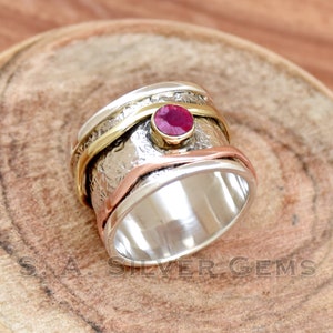Könnte beinhalten: Ein silberner Ring mit strukturiertem Band, mit einem kleinen, runden, roten Edelstein. Der Ring hat gold- und kupferfarbene Bänder um die Mitte. Der Text "S.A. SILVER GEMS" ist sichtbar.