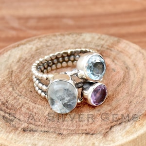 Op de afbeelding: Een zilveren ring met drie edelstenen: een lichtblauwe aquamarijn, een heldere maansteen en een paarse amethist. De ring heeft een geperld bandje.