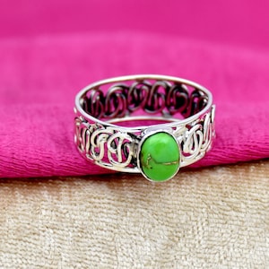 May include: A silver ring with an intricate design and a green gemstone. The ring is set on a pink and beige fabric background.