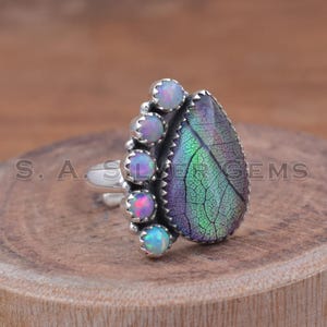 May include: A silver ring featuring a leaf-shaped iridescent gemstone with green, purple, and blue hues. The ring is accented with a row of small, round opals in shades of pink and blue. The ring is set on a wooden surface.