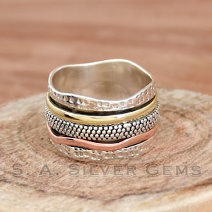 Può includere: Un anello girevole in argento, oro e rame con una superficie testurizzata. L'anello è composto da più bande che girano liberamente.