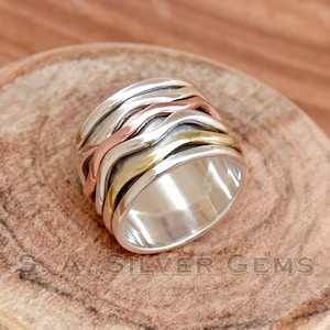 Può includere: Un anello d'argento con tre bande di metallo di diversi colori. Le bande sono intrecciate in un motivo ondulato. L'anello è su una superficie di legno.