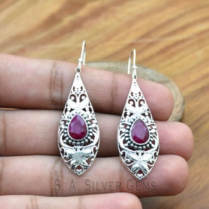 May include: Silver dangle earrings with a teardrop-shaped design. The earrings feature a red gemstone set in a filigree frame with butterfly accents.
