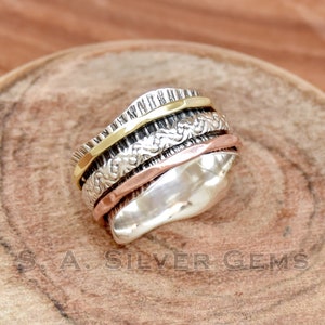 Può includere: Un anello spinner in argento con fasce dorate, ramate e strutturate. L'anello ha un design unico con più fasce rotanti. L'anello è esposto su una superficie di legno. Il testo "S. A. SILVER GEMS" è visibile.