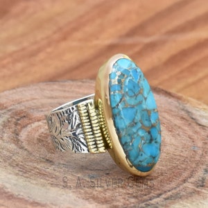 May include: A silver ring with a gold band and a large oval turquoise stone. The stone has a blue and green color and a marbled pattern.