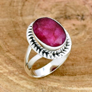 Puede incluir: Un anillo de plata con una gran piedra preciosa de rubí de forma ovalada. El anillo tiene una banda texturizada y detallada.
