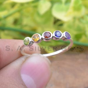 Puede incluir: Un anillo de plata con cinco piedras preciosas de colores: verde, amarillo, rojo, azul y morado. El anillo está sobre un fondo borroso verde y marrón. El texto "S. A. SILVER GEMS" es visible.