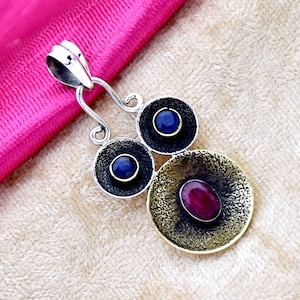 May include: A silver pendant with three round, textured metal circles. The top circle has a blue gemstone, the middle circle has a blue gemstone, and the bottom circle has a red gemstone.