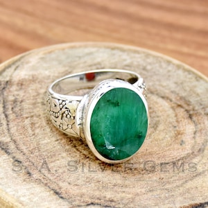 Puede incluir: Un anillo de plata con una esmeralda verde ovalada engastada en un engaste de bisel. La banda del anillo está decorada con diseños florales.