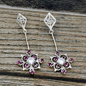 Puede incluir: Un par de pendientes de plata con diseño floral. Cada pendiente tiene un gran cristal transparente rodeado de cristales rosas más pequeños.