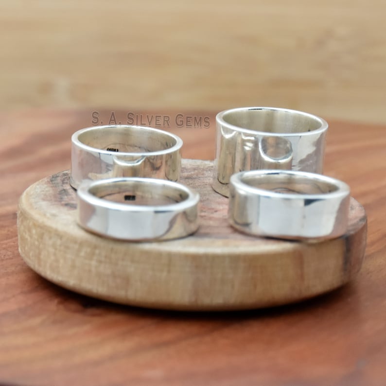 May include: Four silver rings of varying sizes are displayed on a wooden surface. The rings are all smooth and polished, with a slightly rounded edge. The rings are arranged in a semi-circle, with the largest ring at the top and the smallest ring at the bottom. The text "S.A. SILVER GEMS" is visible in the background.