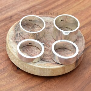 May include: Four silver rings on a wooden surface. The rings are all the same width and have a smooth, polished finish. The rings are stamped with the text "S.A. SILVER GEMS".
