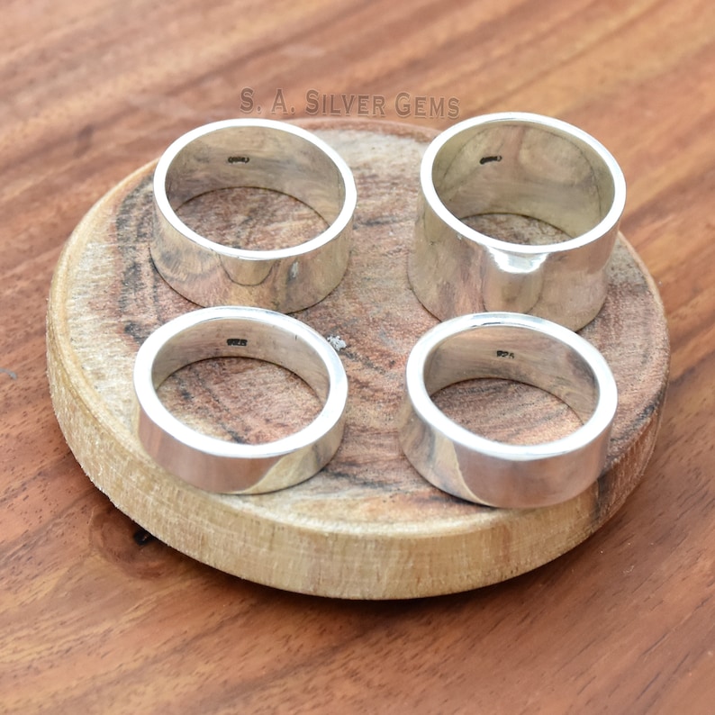 May include: Four silver rings on a wooden surface. The rings are all the same width and have a smooth, polished finish. The rings are stamped with the text "S.A. SILVER GEMS".