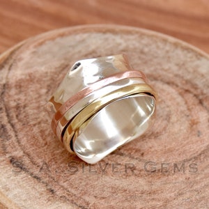 Può includere: Un anello rotante in argento con tre bande di rame e oro. L'anello è su una superficie di legno.
